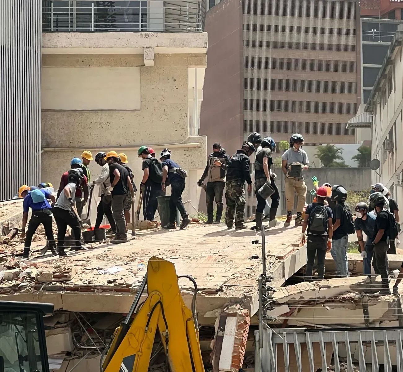 Rescue workers and volunteers in helmets stand on a collapsed concrete structure in Caracas, working together to clear earthquake debris amid high-rise buildings.