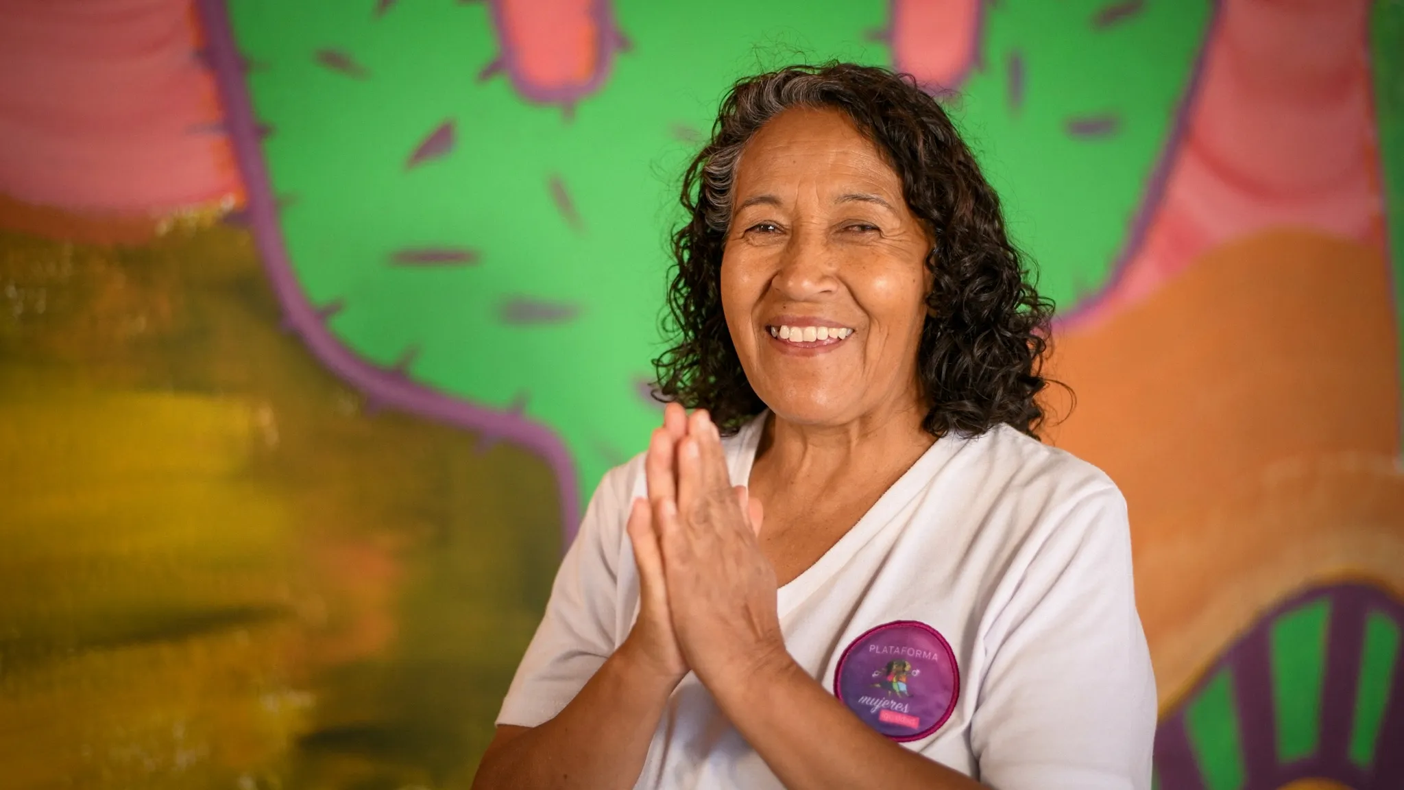A woman with curly, dark hair smiles into the camera with her hands pressed together.