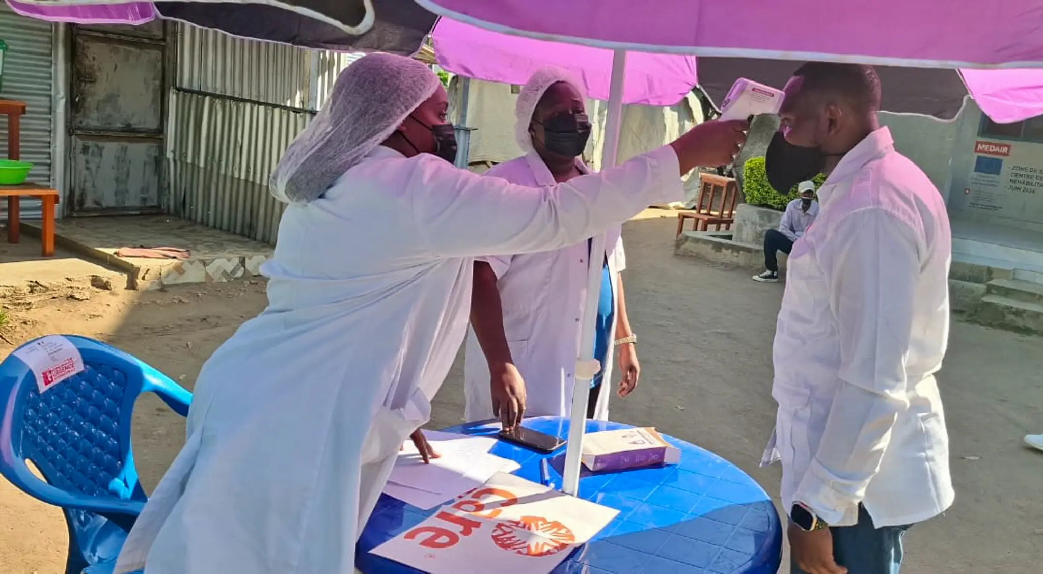 Aid workers at an outside medical clinic taking the temperature of man in white lab coat.