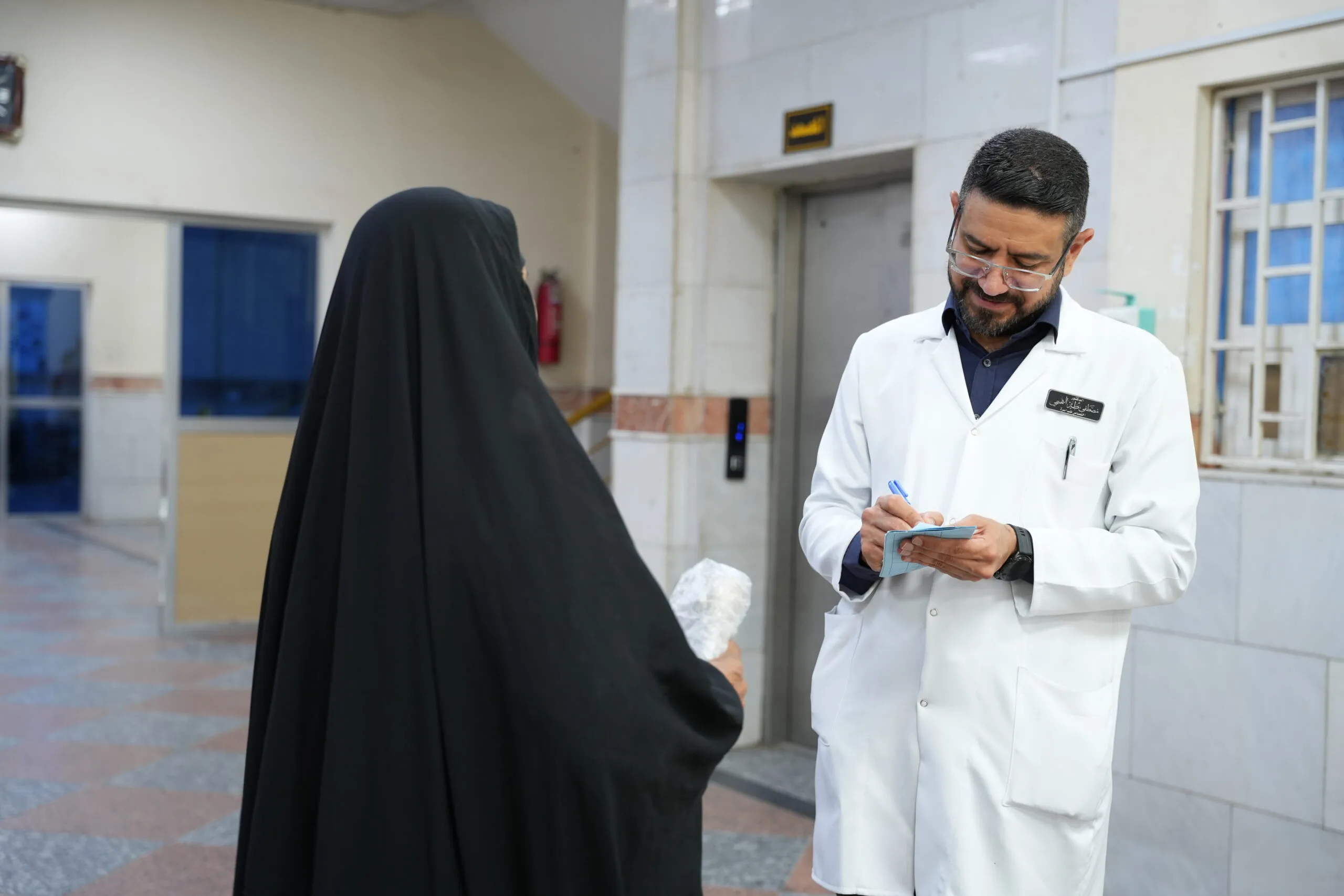 A doctor in a white coat is writing in a notepad and speaking with a woman wearing a black hijab.