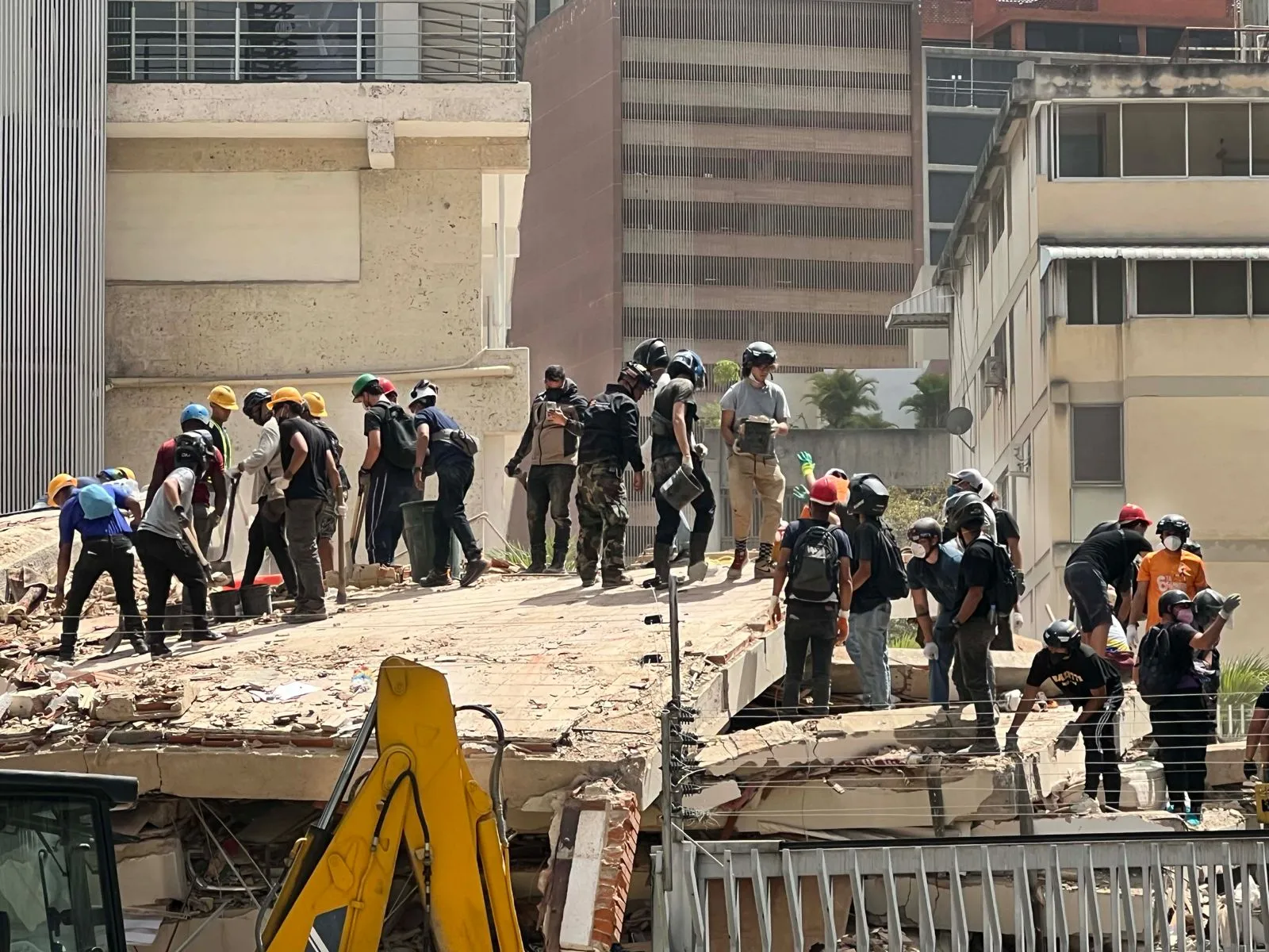 Rescue workers and volunteers in helmets stand on a collapsed concrete structure in Caracas, working together to clear earthquake debris amid high-rise buildings.