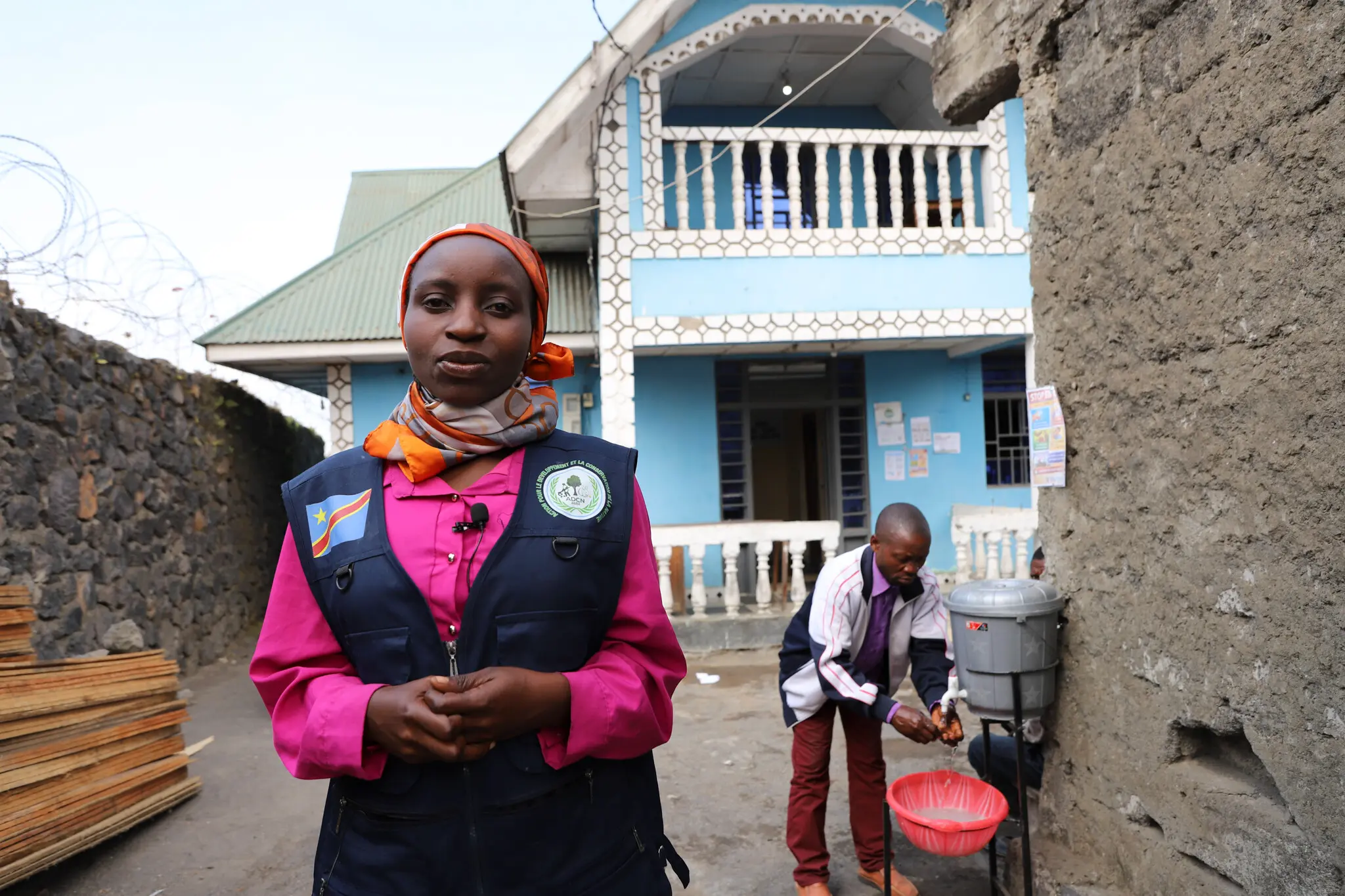 A young woman from the DRC in a vest and headscarf stands in a narrow alleyway, behind her a young man is washing his hands.