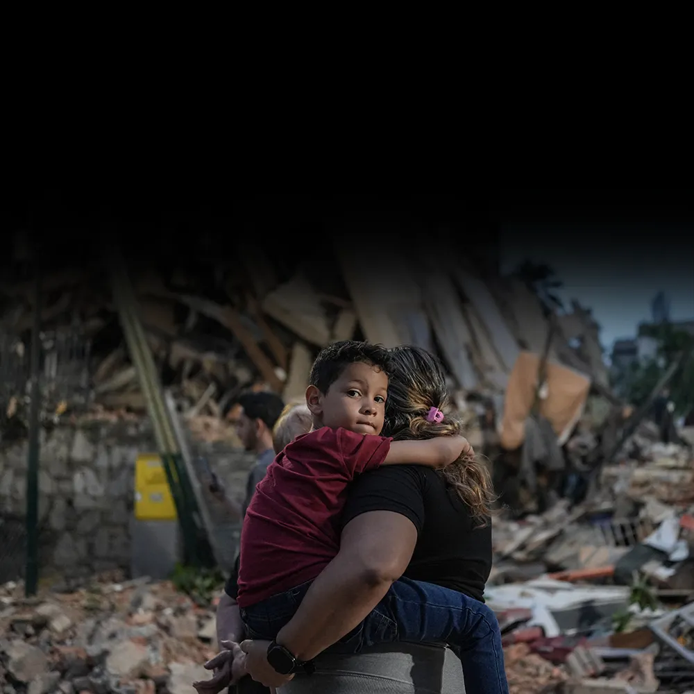 A Venezuelan mother holds a child in her arms. The child looks at the camera while the mother looks at the rubble of a collapsed building.