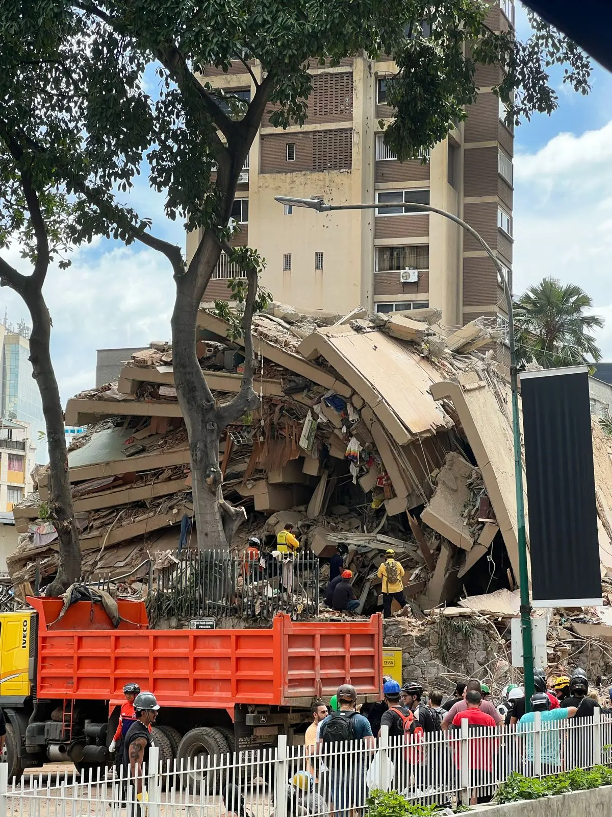 First responders inspect a completely collapsed building reduced to rubble in Caracas, Venezuela, while an orange dump truck and onlookers stand nearby.