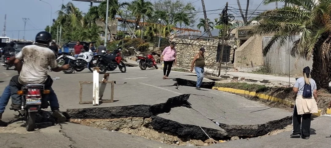 Pedestrians and a motorcyclist navigate a severely cracked and buckled asphalt road in Venezuela following an earthquake.
