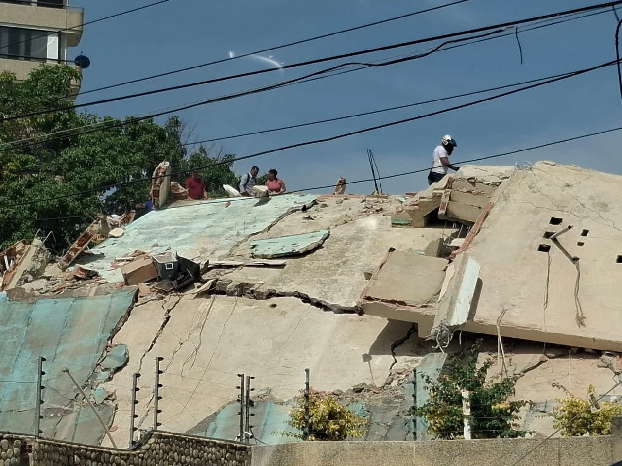 A group of people stands atop a severely buckled and tilted concrete building structure in La Guaira, Venezuela, inspecting the earthquake damage amidst overhead power lines.
