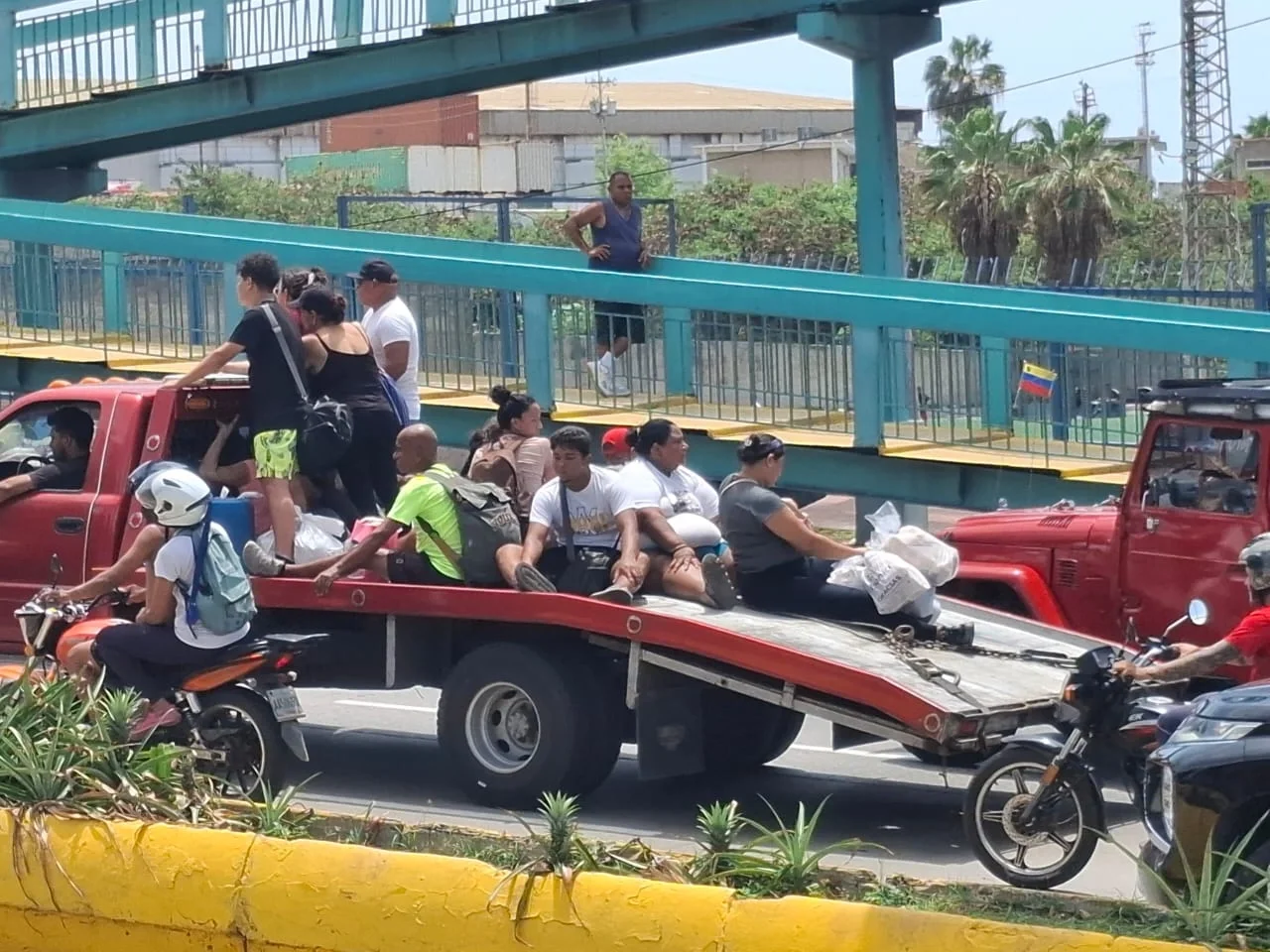 Displaced people carrying bags of belongings ride on the open flatbed of a red truck down a Venezuelan street next to a pedestrian overpass.