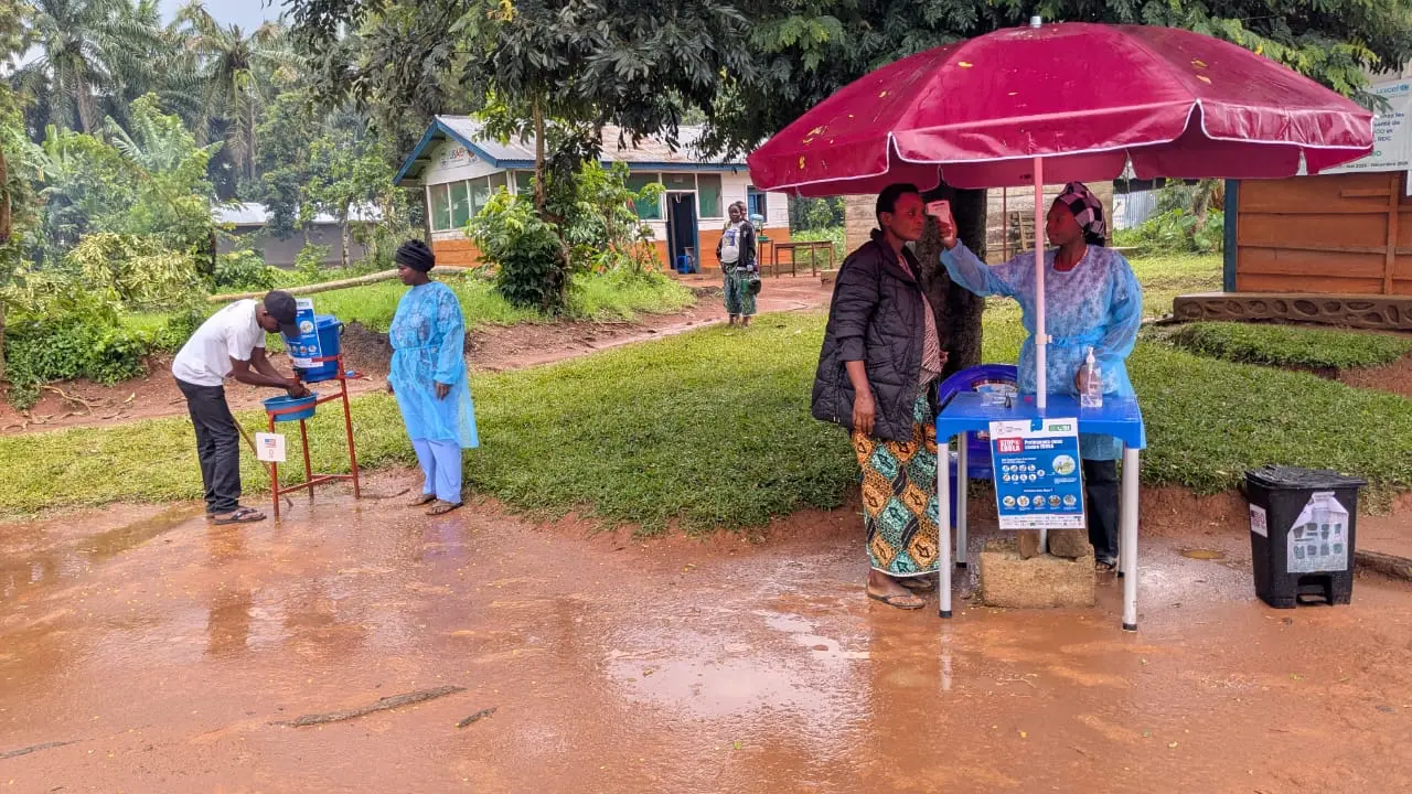 People gathered outside on a rainy day. One person is getting their temperature checked under an umbrella at a makeshift health checkpoint.