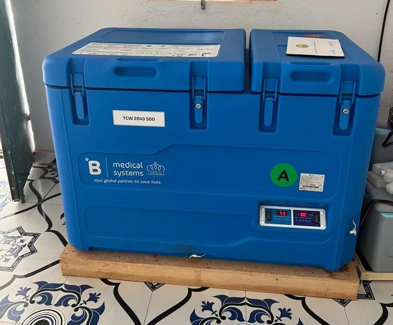 A robust blue solar-powered vaccine refrigerator sits on a wooden platform in a healthcare clinic in Somaliland.