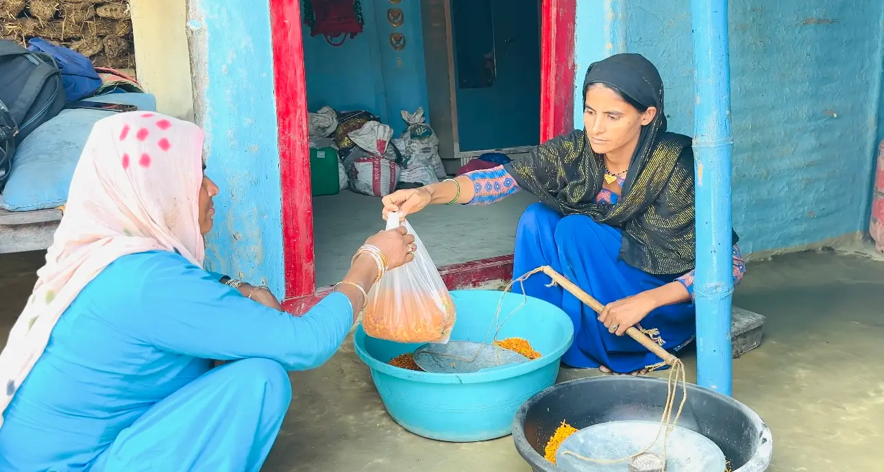 Two women sitting down next to a big bowl at an outdoor market. One woman is handing the other woman a bag of grains.