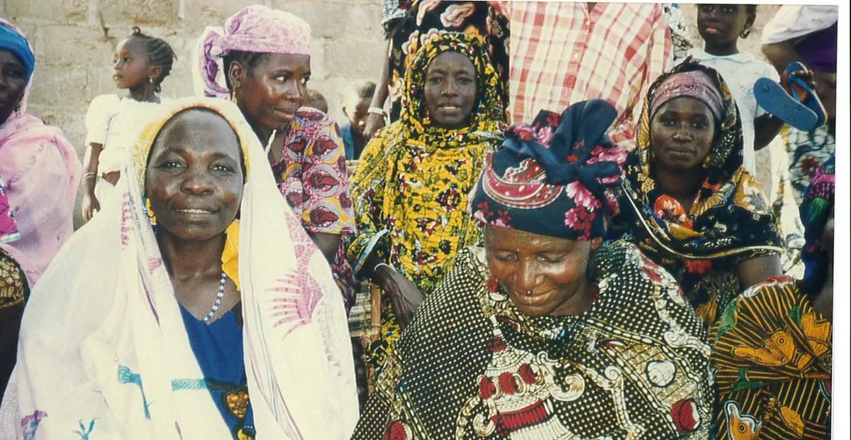 A group of women in colorful headscarves and wraps stand and sit together outside a stone building in a Niger village.