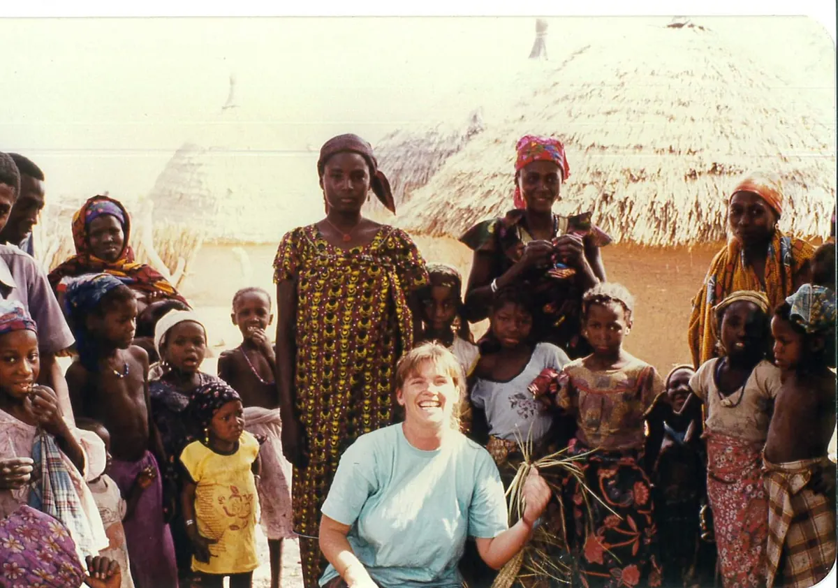 A young woman in a light t-shirt kneels among a group of women and children outside thatched-roof homes.