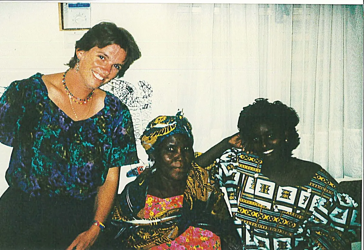 Three women sit together indoors; a woman in a patterned dress is in the middle, flanked by a younger woman and Moira, all smiling.