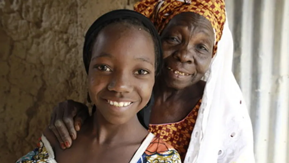 An older woman in a headscarf smiles with her arm around a teenage girl, standing against a mud wall.