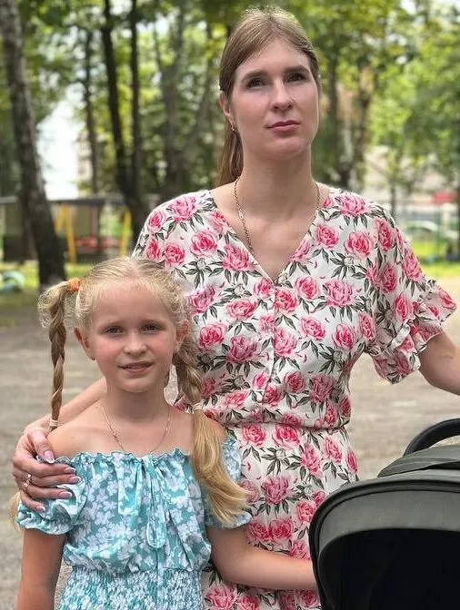A mother in Ukraine stands outside a park with her young daughter and toddler son sleeping in stroller.