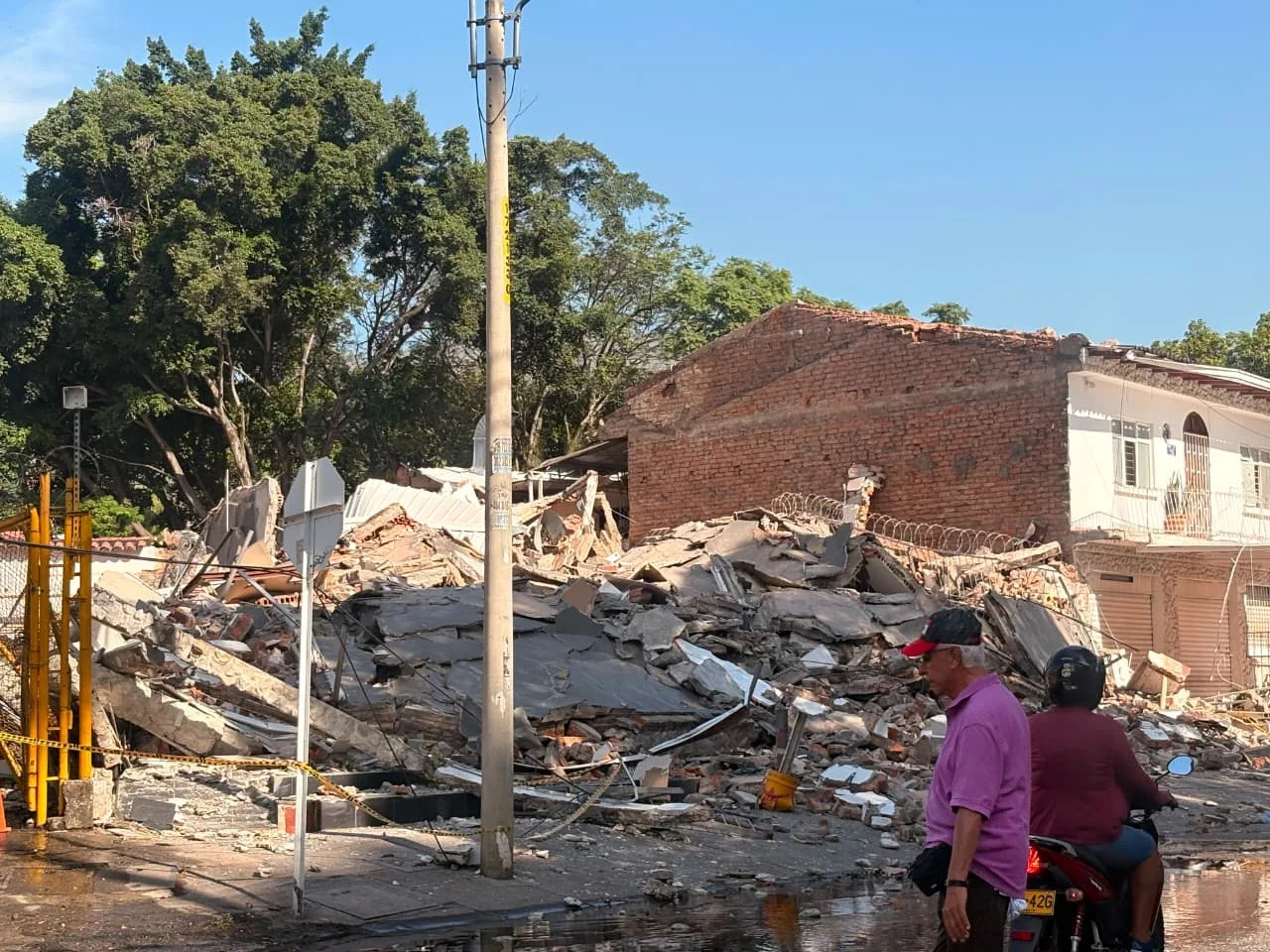 A completely collapsed brick building reduced to a pile of rubble and concrete debris along a street, with bystanders and a motorcyclist nearby showing the aftermath of a Colombia earthquake.