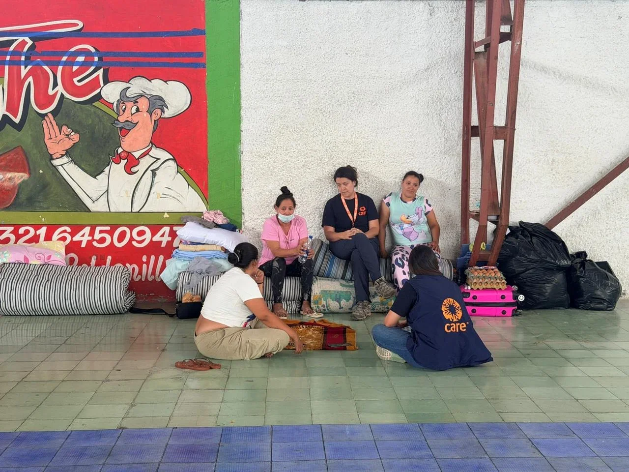 Aid workers wearing CARE vests speak with displaced people sitting on mattresses with their belongings in a temporary shelter after the Colombia earthquake.