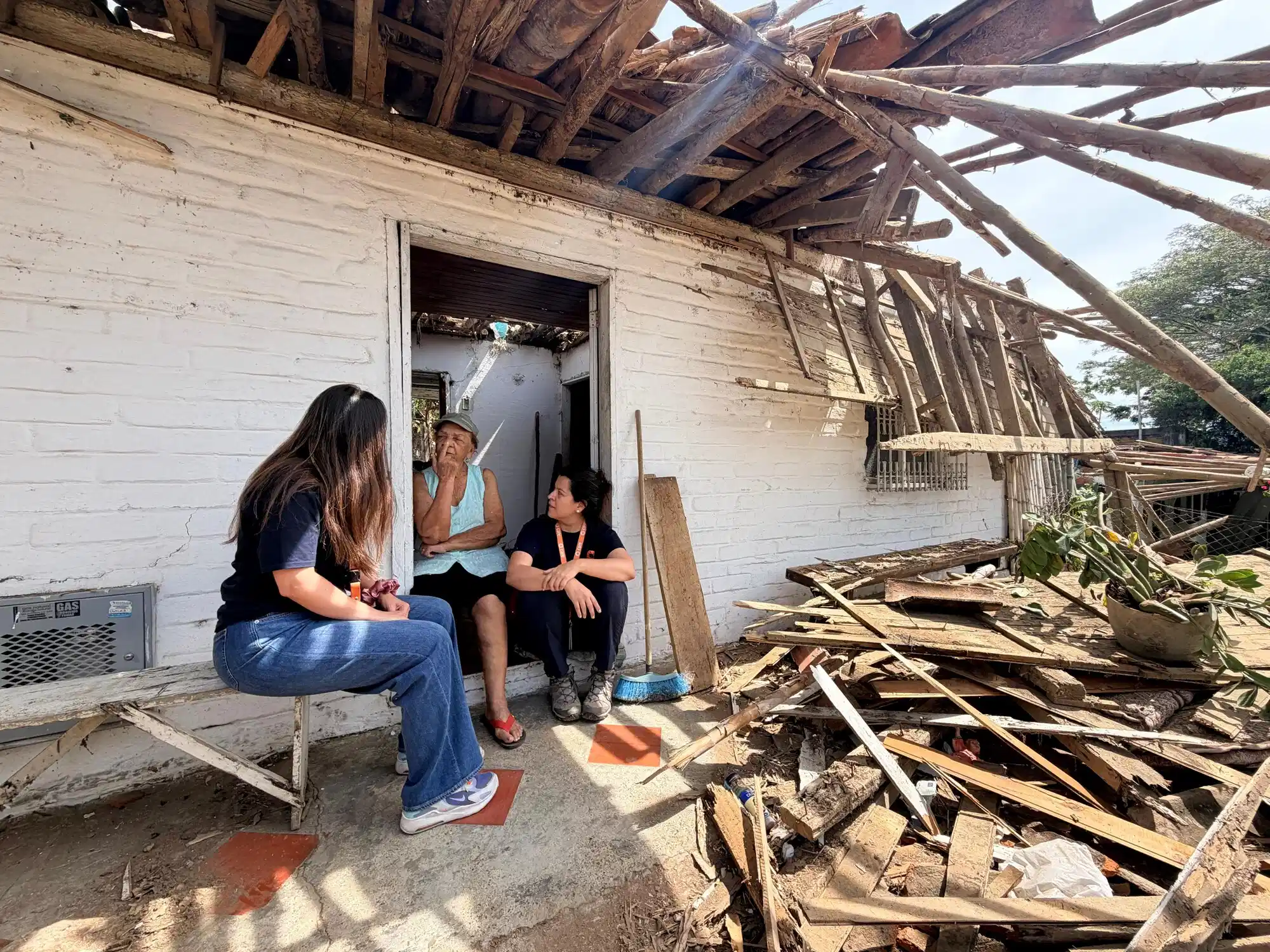 An elderly woman and two aid workers sit at the doorway of a house with a severely damaged, collapsed wooden roof and surrounding debris, illustrating the aftermath of a Colombia earthquake.