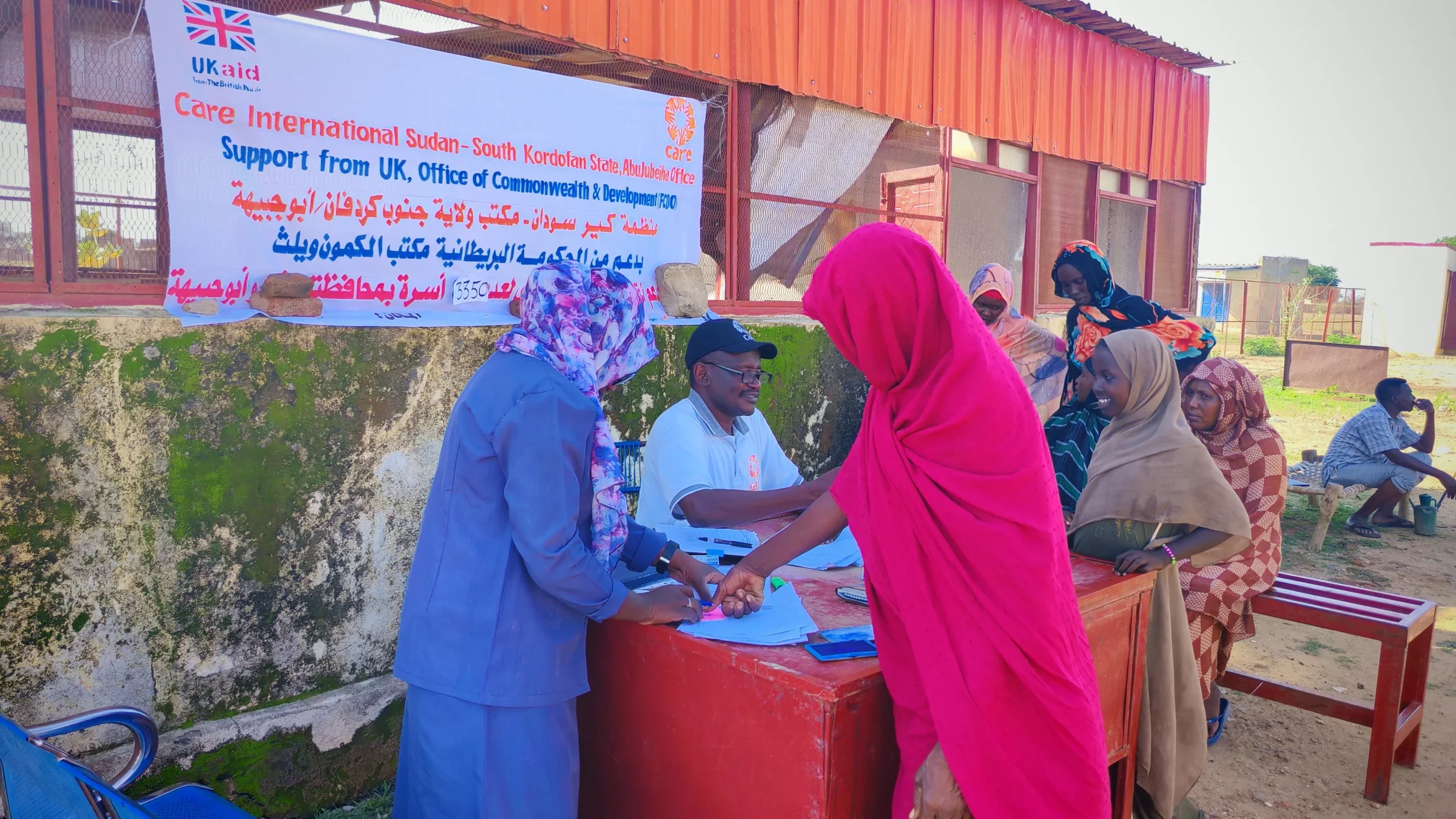 Hawa, seen from behind to protect her identity, gives her fingerprint to register for cash assistance at a desk in Abu Jubayhah, South Kordofan, while other community members wait nearby.