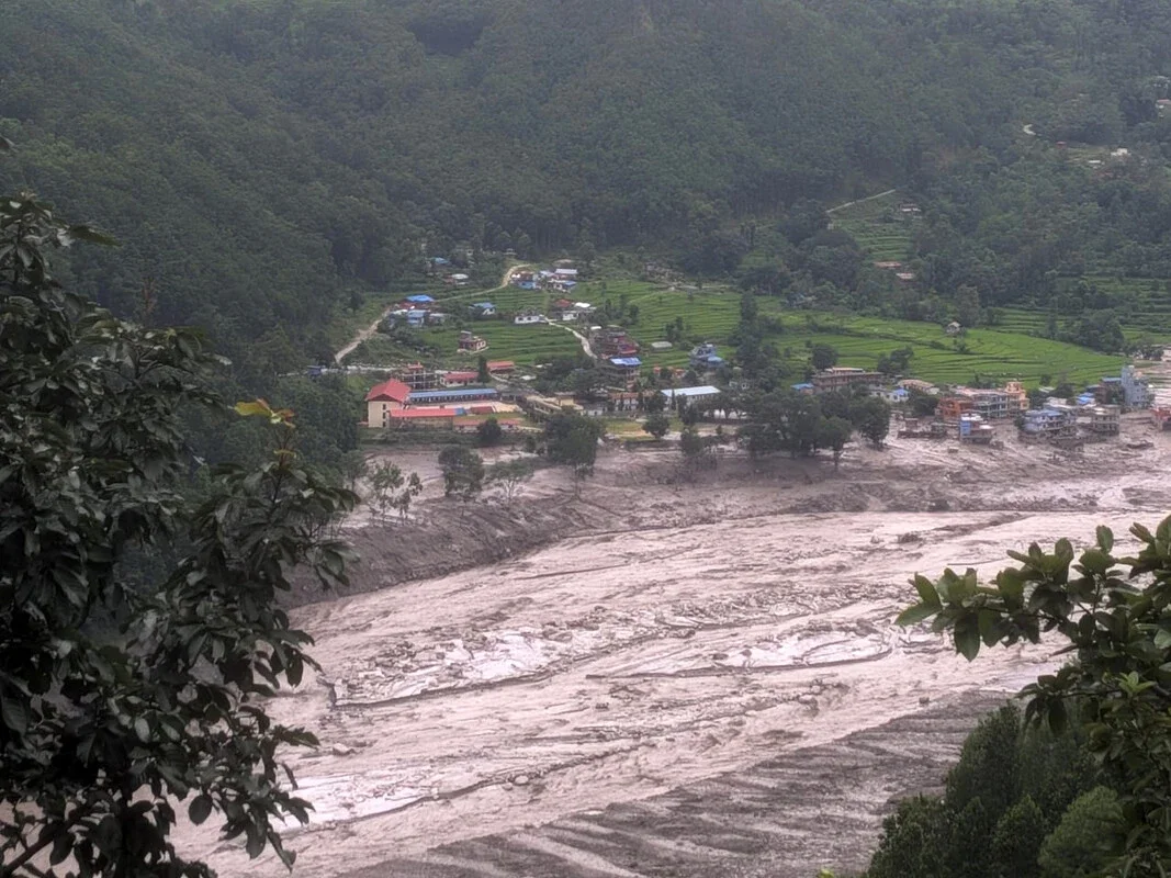 Aerial view of a wide, devastated riverbed filled with mud and debris flowing through a green Himalayan valley, with intact red-roofed buildings and green fields on the hillside above.