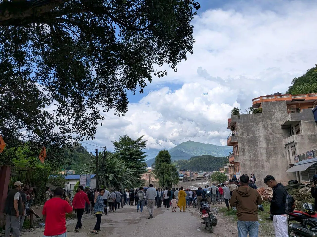 Crowds of people walk along a paved road lined with trees and concrete buildings toward a wide expanse of grey flood debris in the distance, with green hills and clouds beyond.