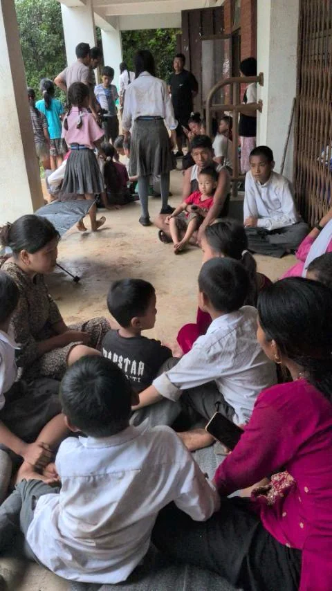 A crowded indoor temporary shelter with dozens of children and adults sitting on the floor and standing in a concrete building with minimal furnishings.