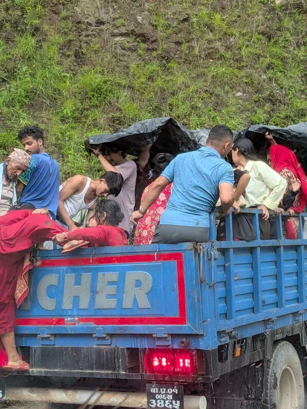 A blue and red cargo truck filled with people holding their belongings as they are transported away from the disaster zone, with mountains visible in the background.