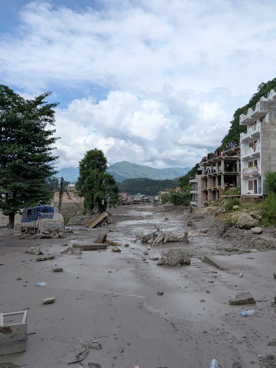 A wide channel of grey mud and rubble runs between damaged buildings in a Rasuwa settlement, with a truck and other vehicles stranded in the debris and people watching from the balconies of a partly built concrete structure.
