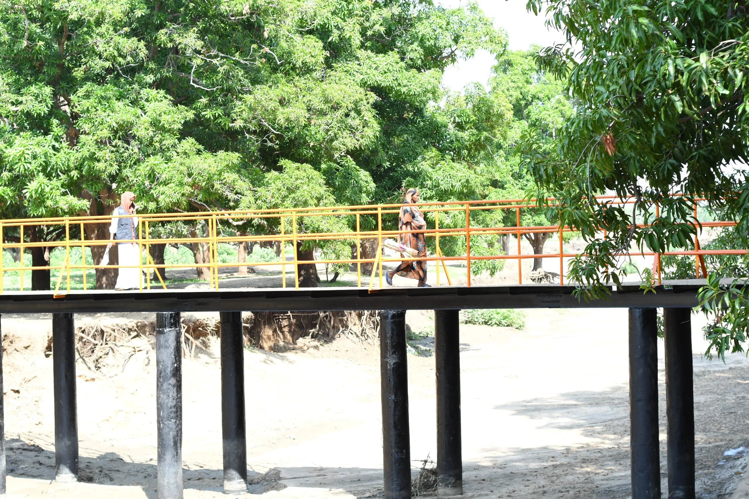 People walking across a newly constructed footbridge with bright yellow and orange handrails, built over a dry riverbed in Alabassiya.