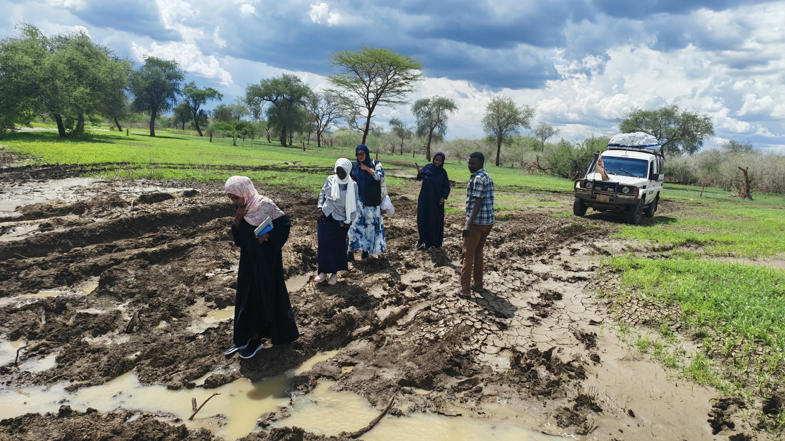 Four female CARE Sudan team members and a male colleague walk on foot through deep mud toward their vehicle under stormy skies while traveling to deliver cash assistance in South Kordofan.