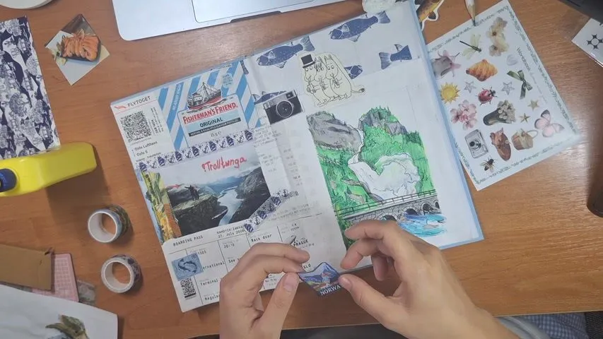 Svitlana’s hands arrange paper clippings and illustrations in an open scrapbook on a table.