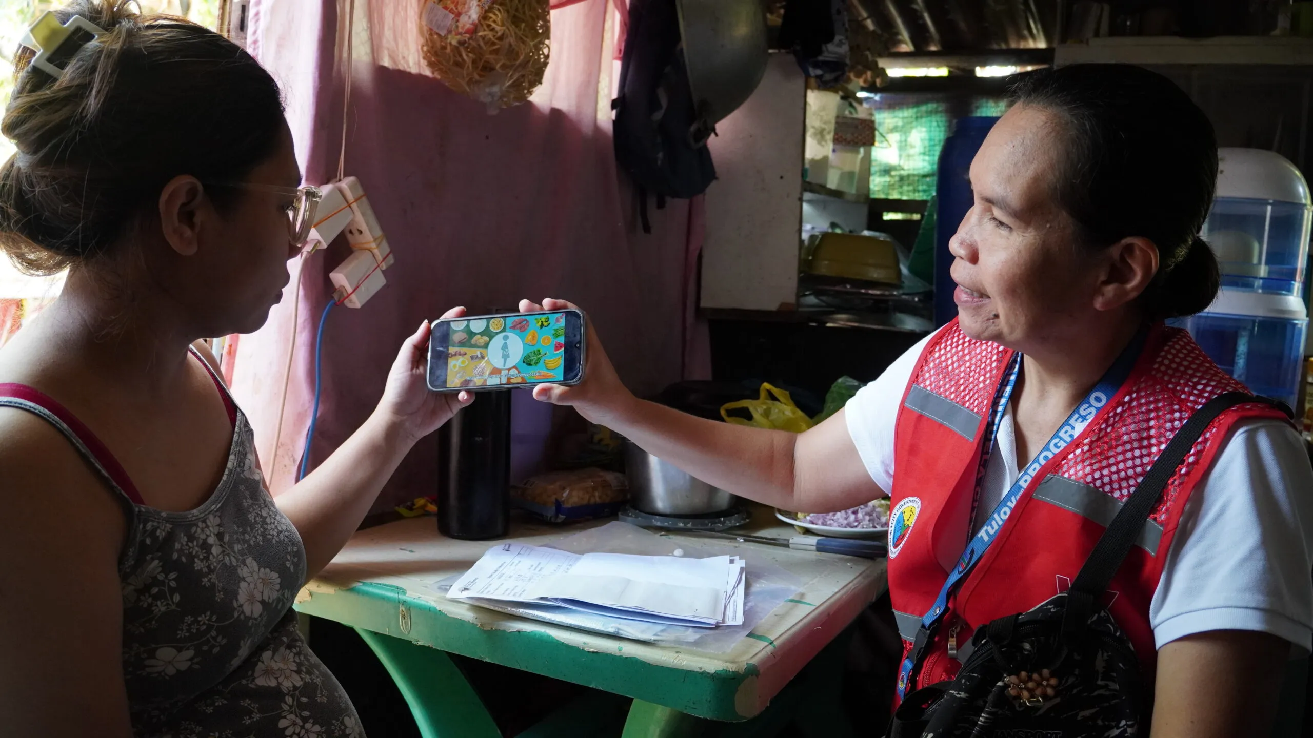 A woman in a medical vest is showing a woman sitting next to her medical advice on a phone.