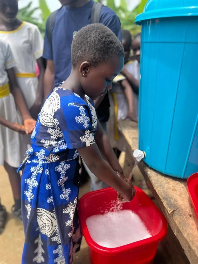 A child washing their hands with water running from barrel into soapy bucket.