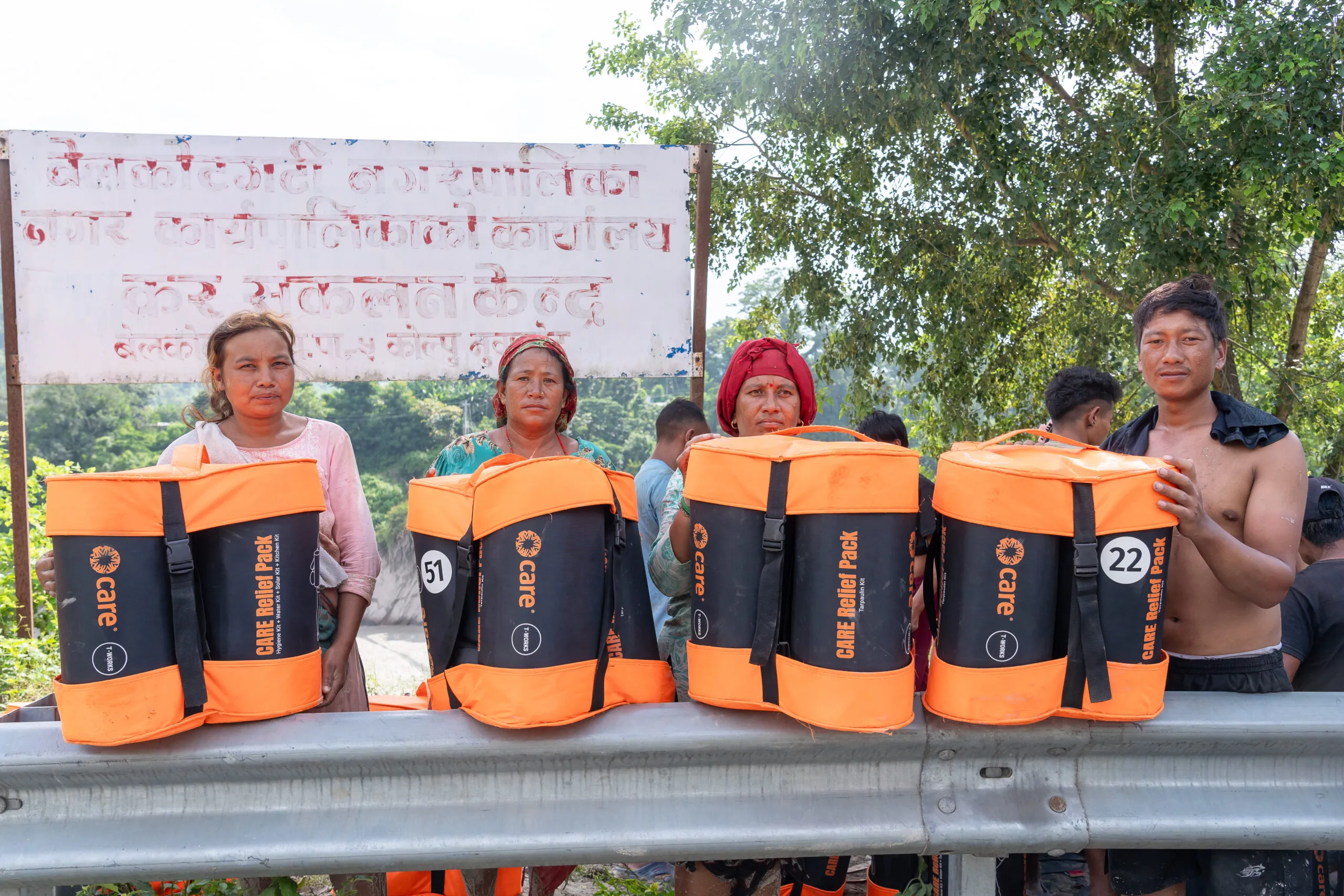 Four women and a young man stand along a roadside guardrail in Nepal, each holding an orange-and-black CARE Relief Pack from a CARE PACKAGE for Emergencies kit, after receiving support for their flood-affected families.
