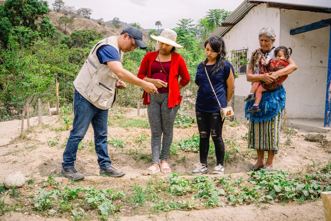 A CARE staff member stands by a small row of crops and talks with three women, one of whom is holding a baby.
