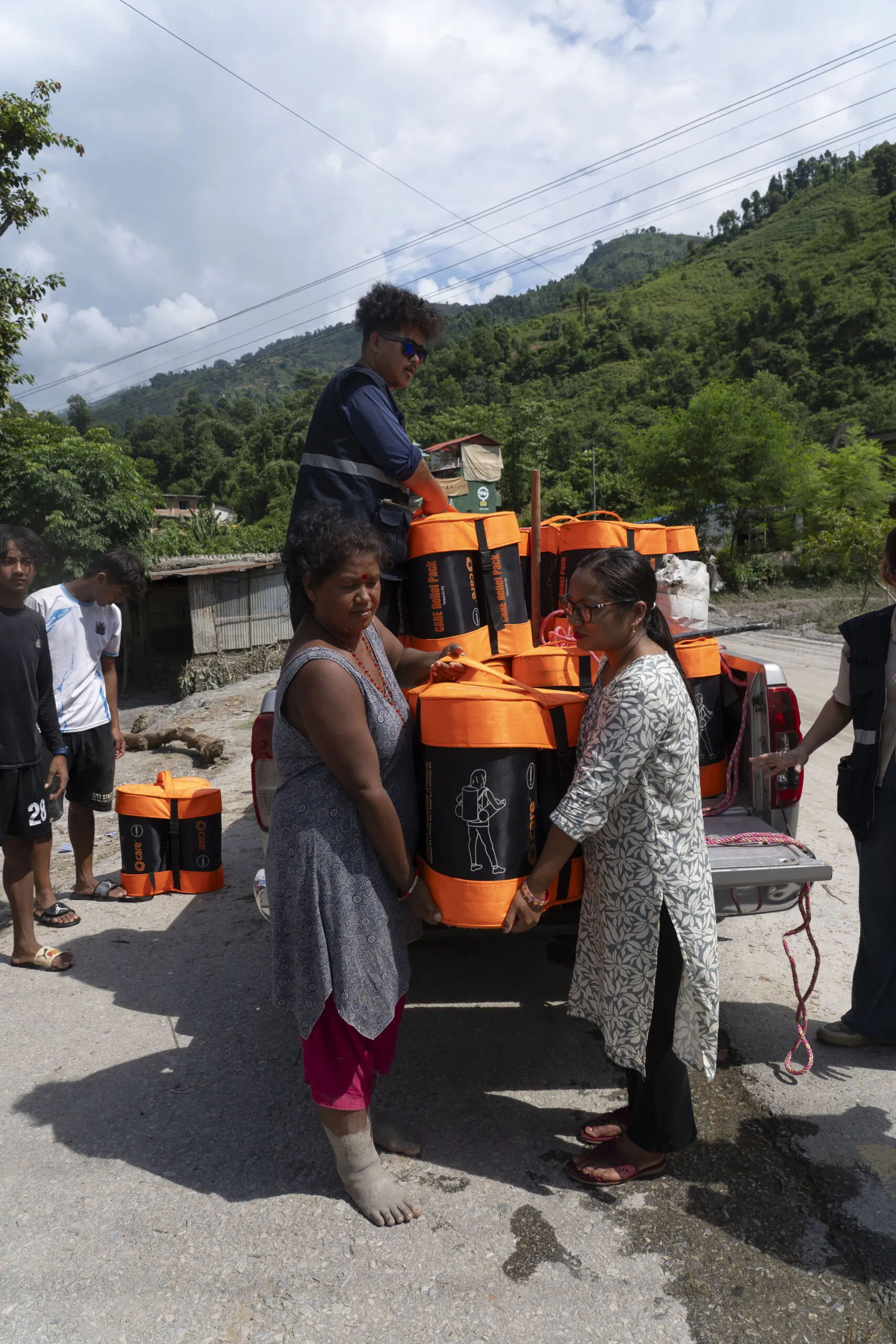 Women receive orange-and-black CARE PACKAGE for Emergencies kits from the back of a truck at a flood distribution site in Belkotgadhi, Nepal.