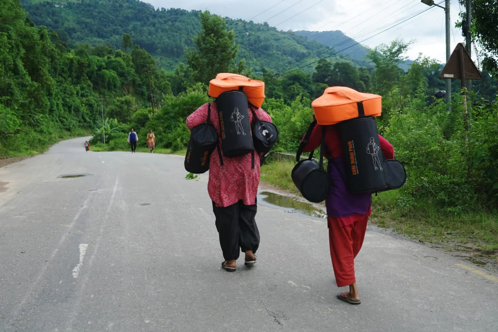 Women walk down a rural road, each carrying a large orange-and-black CARE relief backpack. 