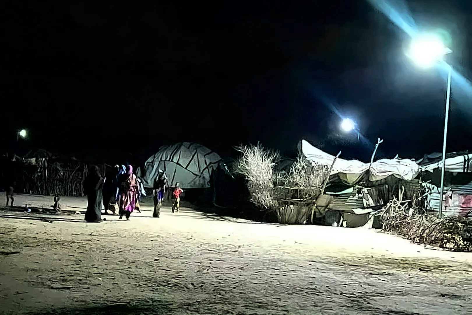 Women and children walk at night, lit by three streetlights above tarpaulin shelters.
