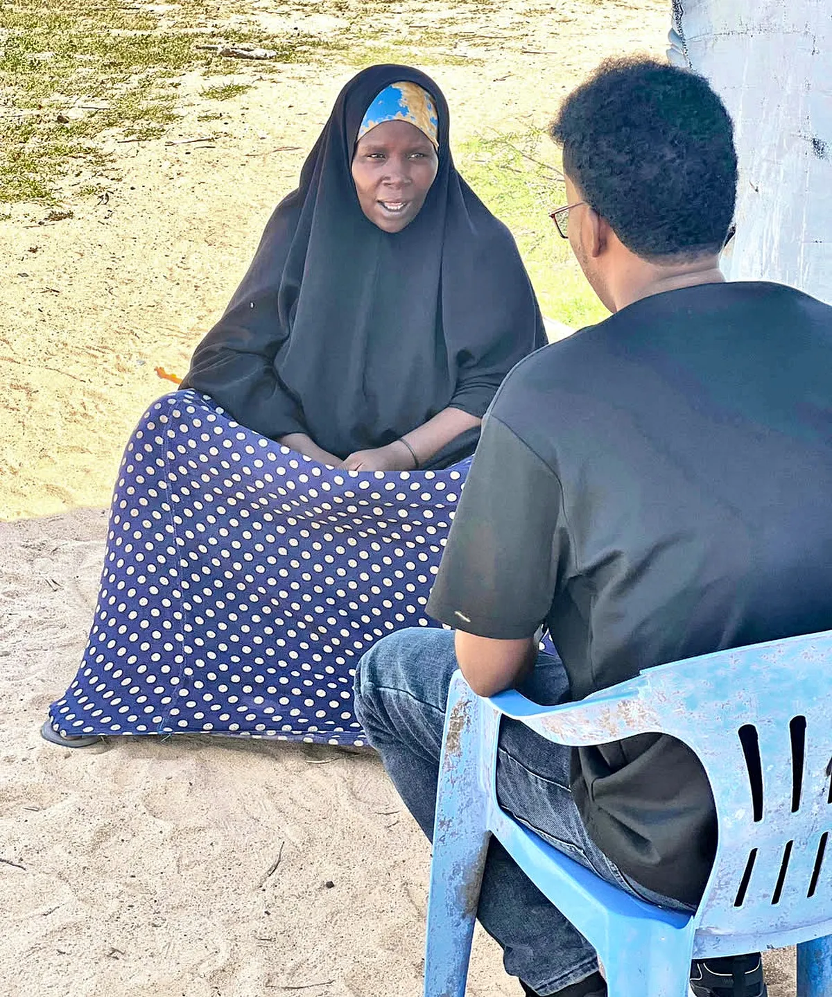 A woman in a dark hijab sits on a patterned mat, speaking to a man seated opposite her in a plastic chair.