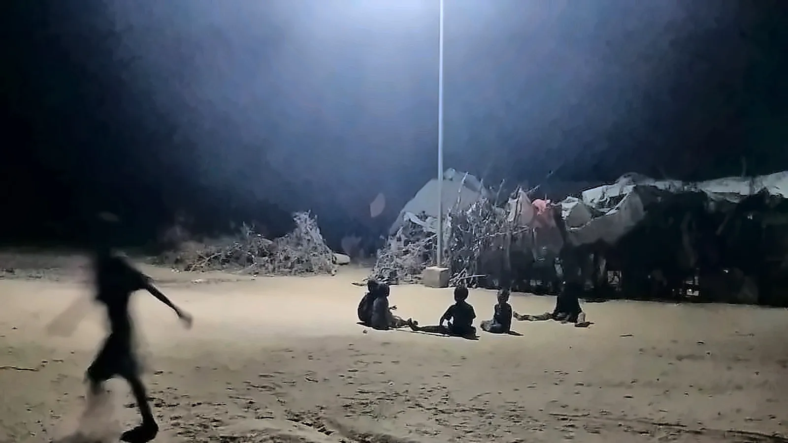 Children sit together on the ground in the pool of light from a streetlight. Another child runs past in the foreground.