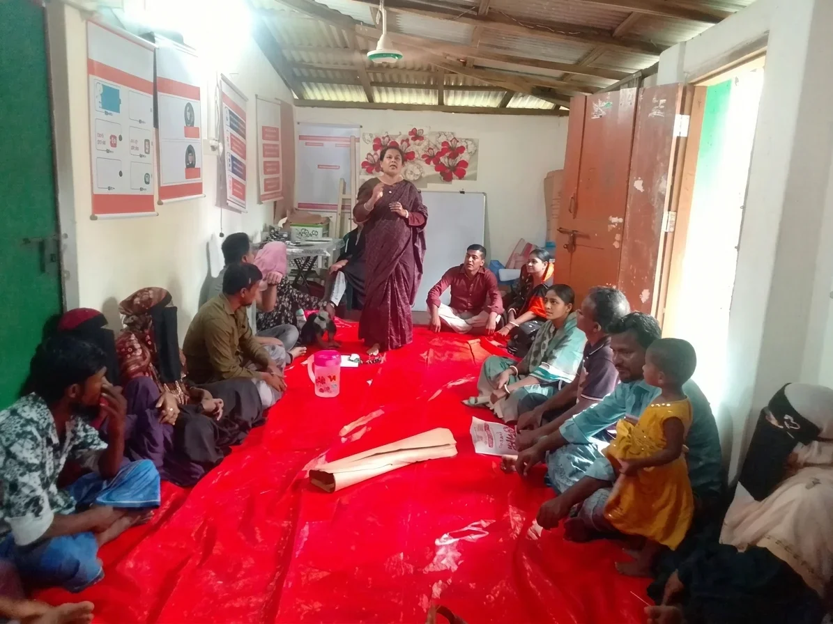 A Bangladeshi woman in workshop settings stands and speaks to a mixed group of men, women, and a child seated on a red tarp indoors.