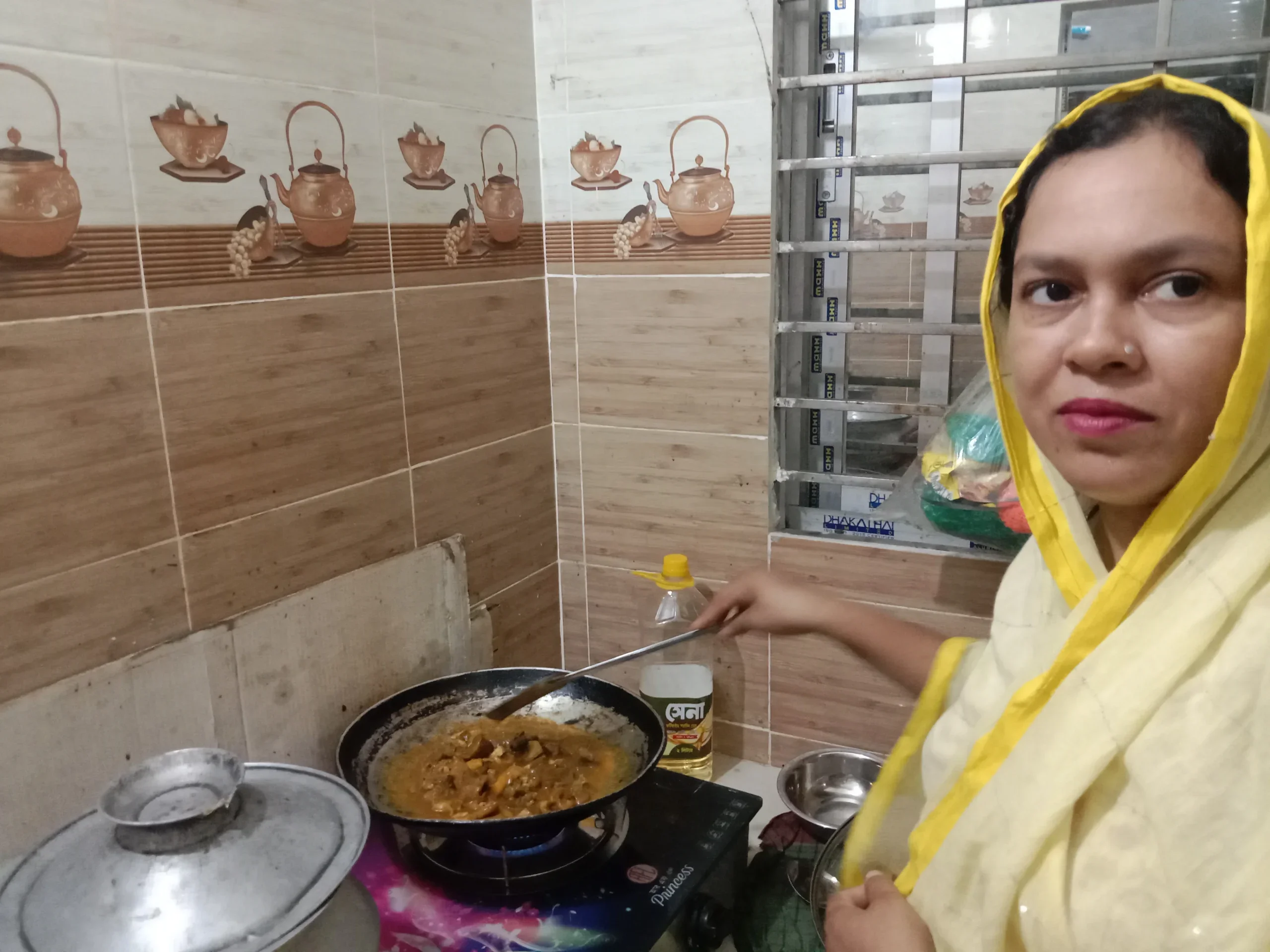 A Bangladeshi woman in a yellow headscarf stirs a pan of curry on a gas stove in a home kitchen.