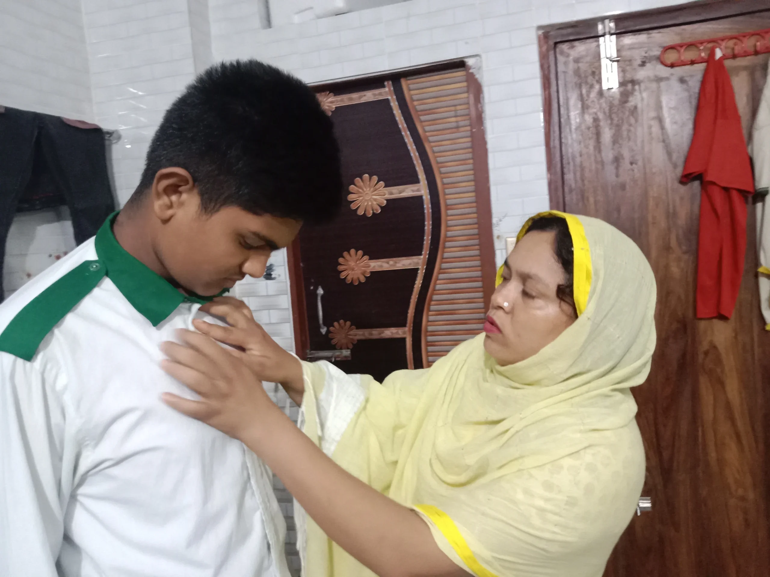 A Bangladeshi woman adjusts the collar of a teenage boy's white school uniform in a home.