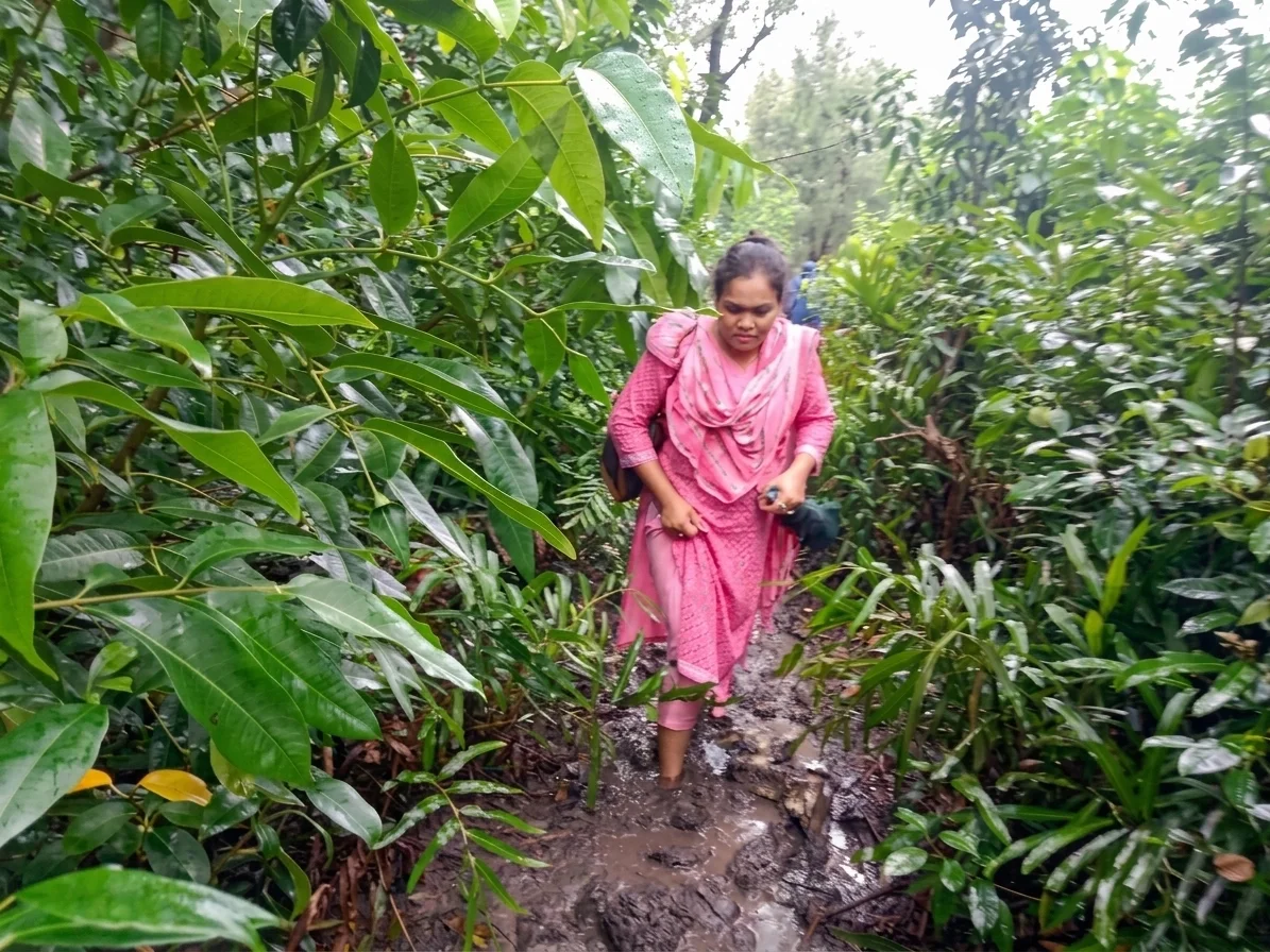 A Bangladeshi woman in a pink salwar kameez wades through muddy, water-logged terrain surrounded by dense green vegetation.