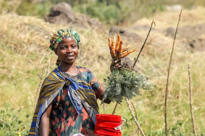 A refugee woman from Nakivale settlement in Uganda in a patterned dress and headwrap speaks expressively while standing in front of rows of green cabbages, representing refugees in Uganda building self-reliance.