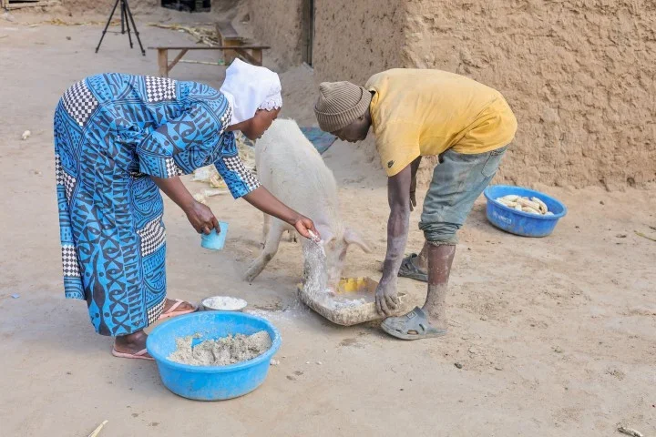 A refugee woman and man from Nakivale settlement in Uganda bend together over a trough to feed a pig outside their home, illustrating the shared livelihood assets built through CARE's EFASeR self-reliance program.