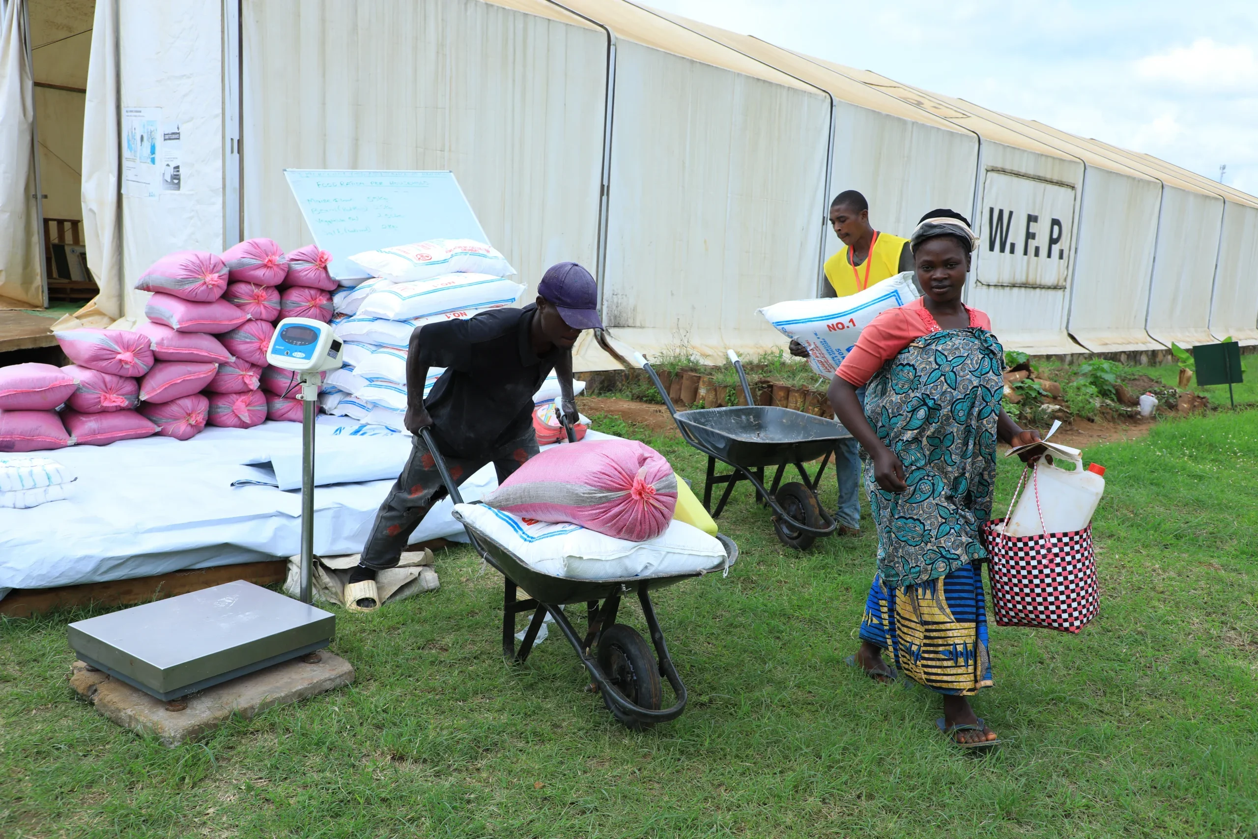 A refugee woman from Nakivale settlement in Uganda walks away from a WFP food distribution point carrying supplies, while workers load sacks of maize and beans onto wheelbarrows outside a large tent marked 