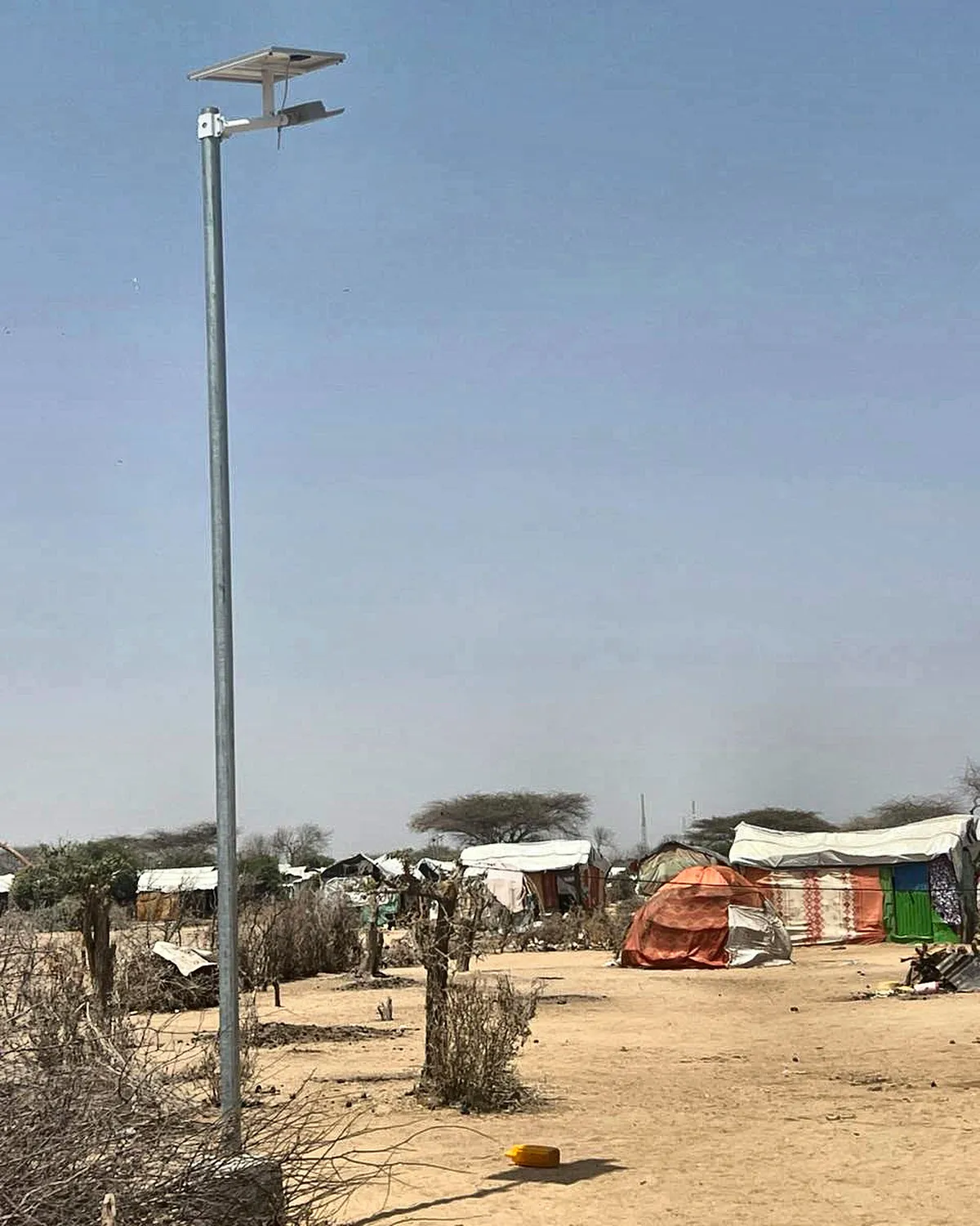 A tall streetlight with a solar panel and lamp at the top stands in sandy ground in front of tarpaulin shelters.