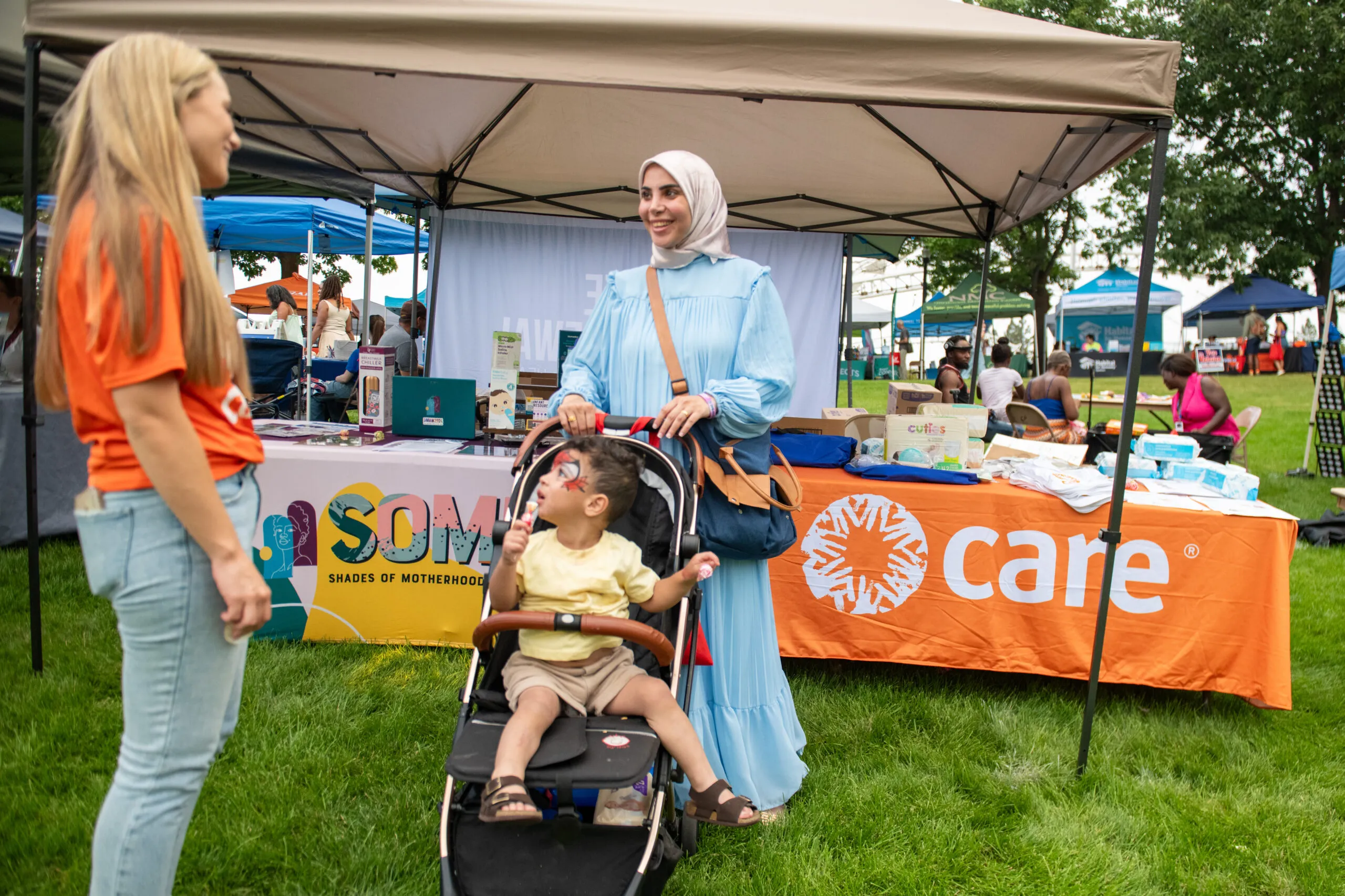 A woman in a CARE shirt speaks with a mother who is standing by her baby in a stroller.