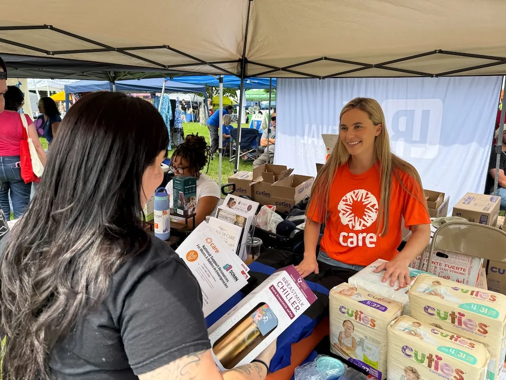 A smiling woman in an orange CARE T-shirt stands behind a table at an outdoor community event, talking with a woman who is holding a breastmilk chiller box. The table is stocked with boxes of Cuties diapers and tip sheets for moms.